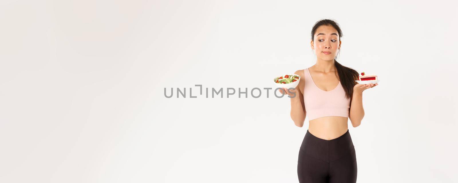 Active lifestyle, fitness and wellbeing concept. Indecisive cute asian slim girl in workout outfit, holding healthy salad and cake, hesitating, need stay on diet, losing weight, white background.