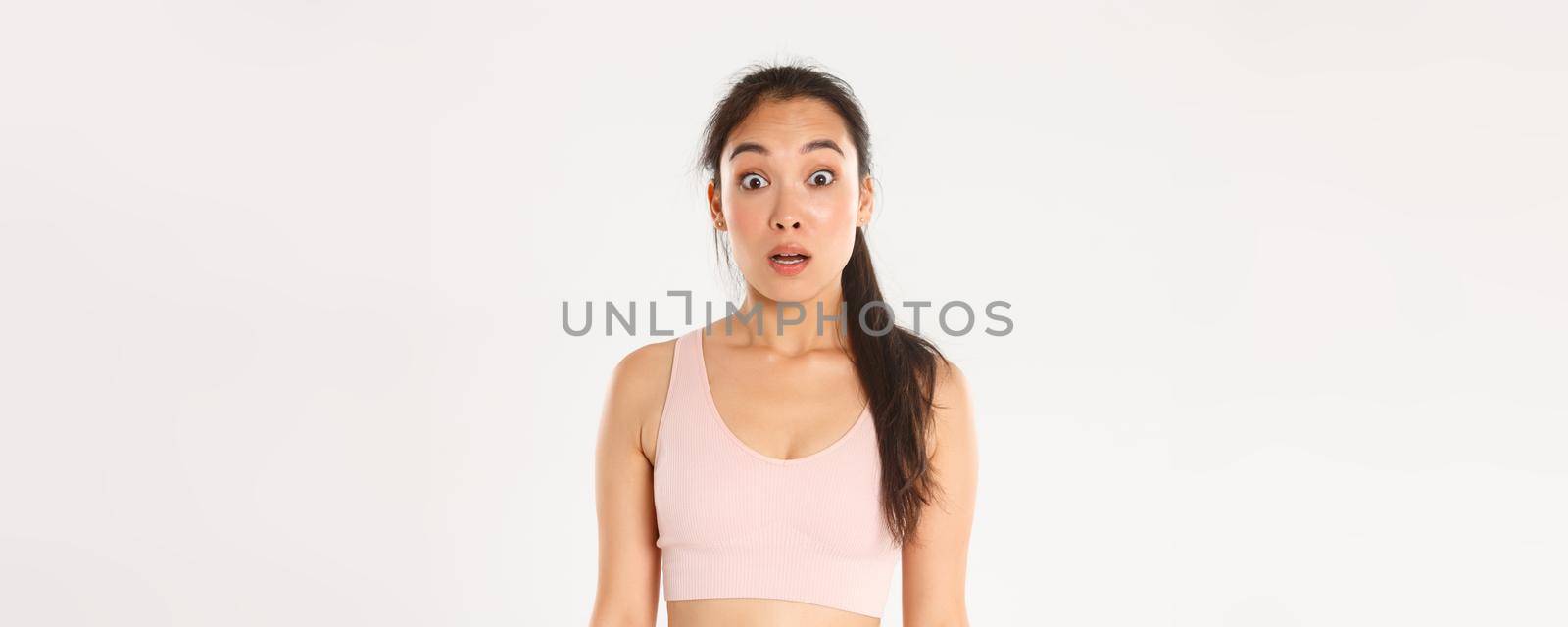 Sport, wellbeing and active lifestyle concept. Close-up of surprised and startled asian fitness girl, sportswoman in sportsbra looking confused with dropped jaw, hear news, white background by Benzoix