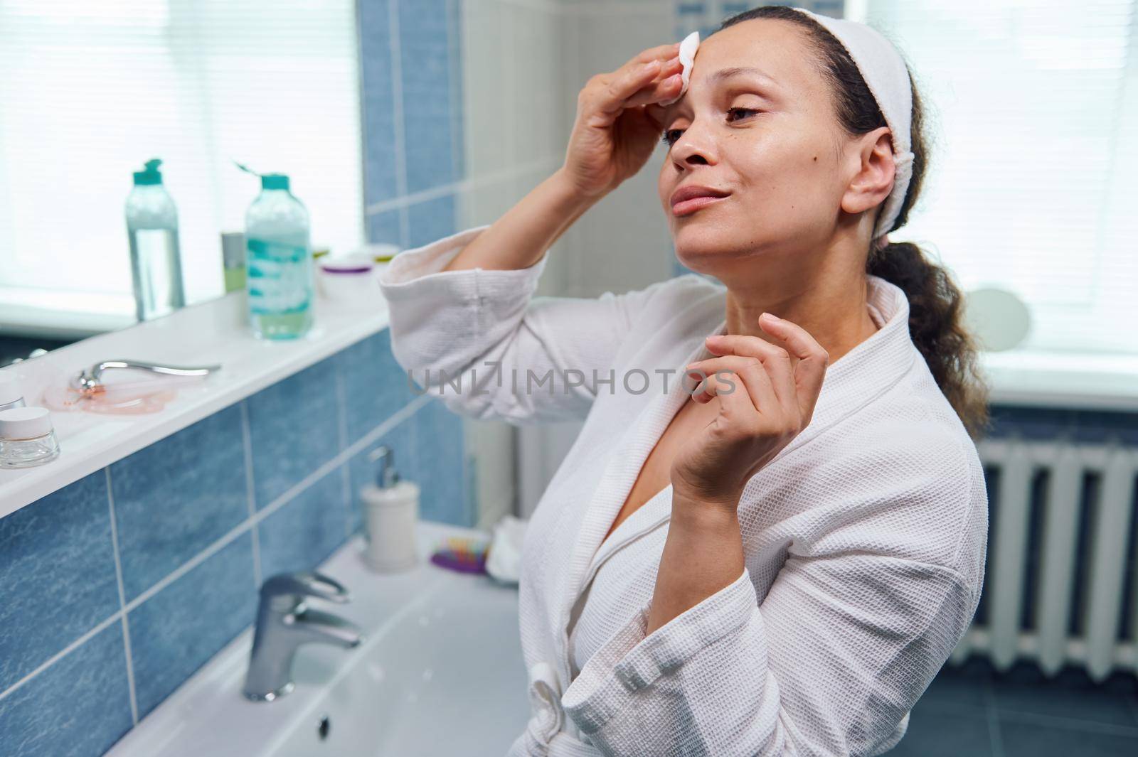 Natural beauty African American woman in white bathrobe, using cleanser cosmetic lotion and cotton pad, removes makeup, enjoys skin care procedures, looking at her reflection in the bathroom mirror