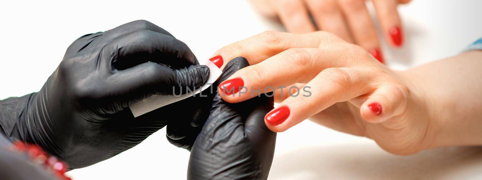 Closeup of manicure master in black gloves wiping the female red nails with a paper napkin in the beauty salon