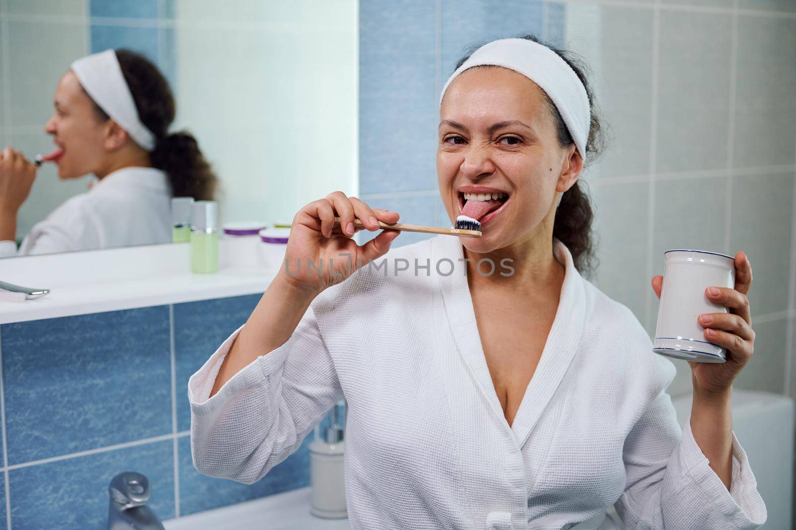 Joyful multi-ethnic woman wearing a white bathroom, standing against a bathroom mirror, tasting a toothpaste with her tongue, smiles at the camera while taking care of her oral hygiene. Dental health