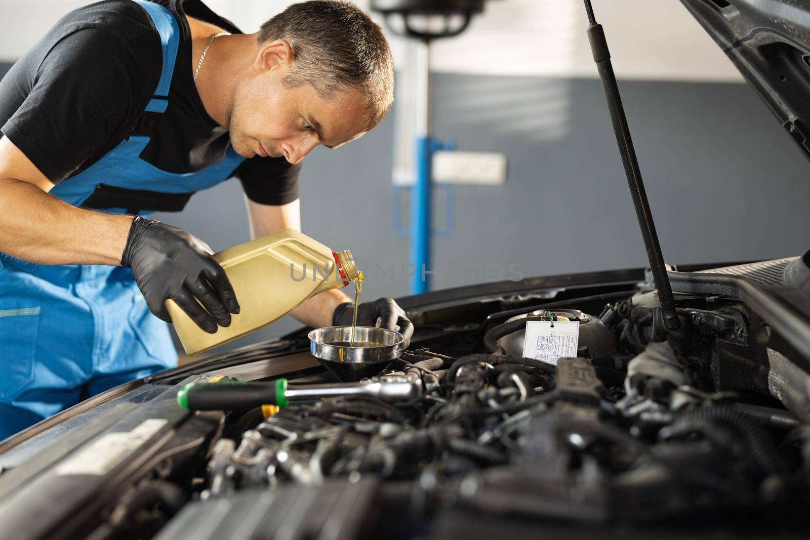 The mechanic is pouring oil into the engine. Pouring fresh oil to car engine, oil change to auto. Caucasian man in blue overalls pouring oil from plastic container by uflypro