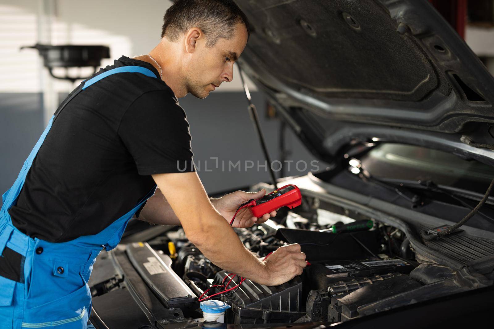 Professional car mechanic check battery voltage with electric multimeter. Automobile diagnosis. Car mechanic repairer looks for engine failure on diagnostics equipment in vehicle service workshop.