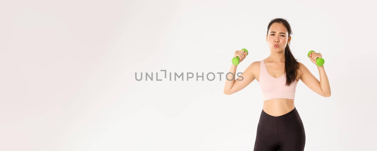 Fitness, healthy lifestyle and wellbeing concept. Portrait of tired asian girl in sportswear, looking exhausted during workout, exercise at home with online coach, lifting dumbbells, white background.