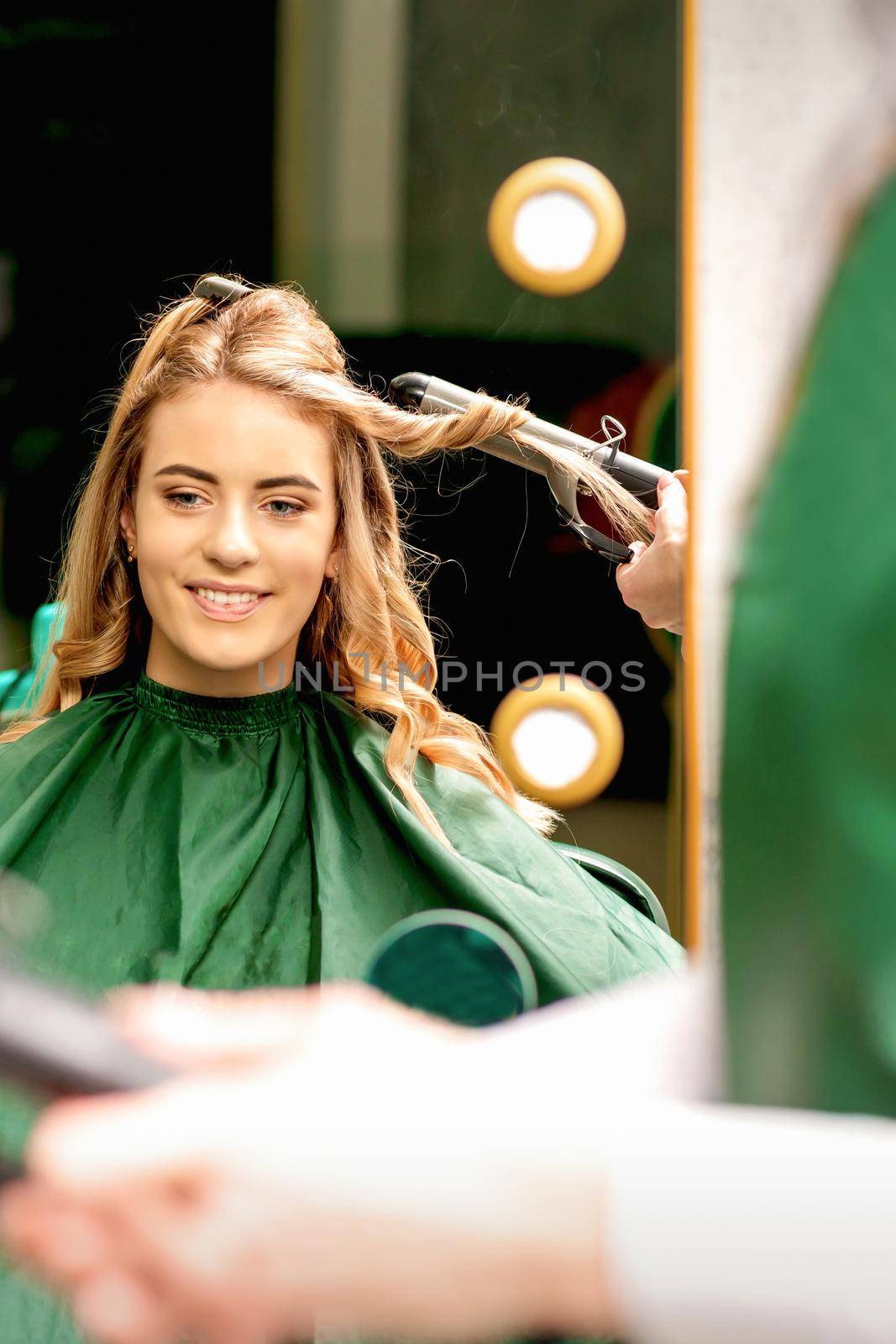 Hairdresser using curling tongs curls long brown hair on the young caucasian girl in a beauty salon
