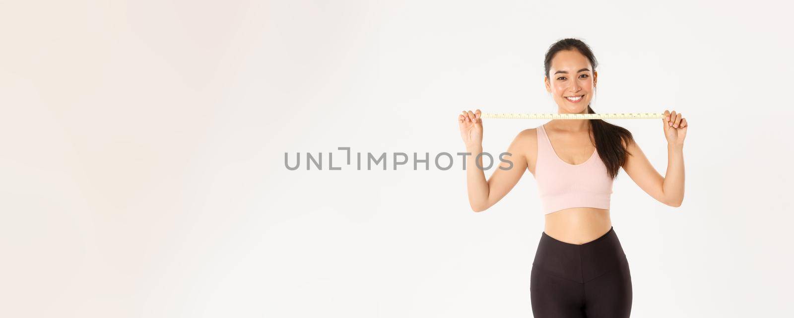 Fitness, healthy lifestyle and wellbeing concept. Portrait of smiling asian brunette sports girl, female athelte in activewear showing measuring tape, losing weight with exercises, white background by Benzoix