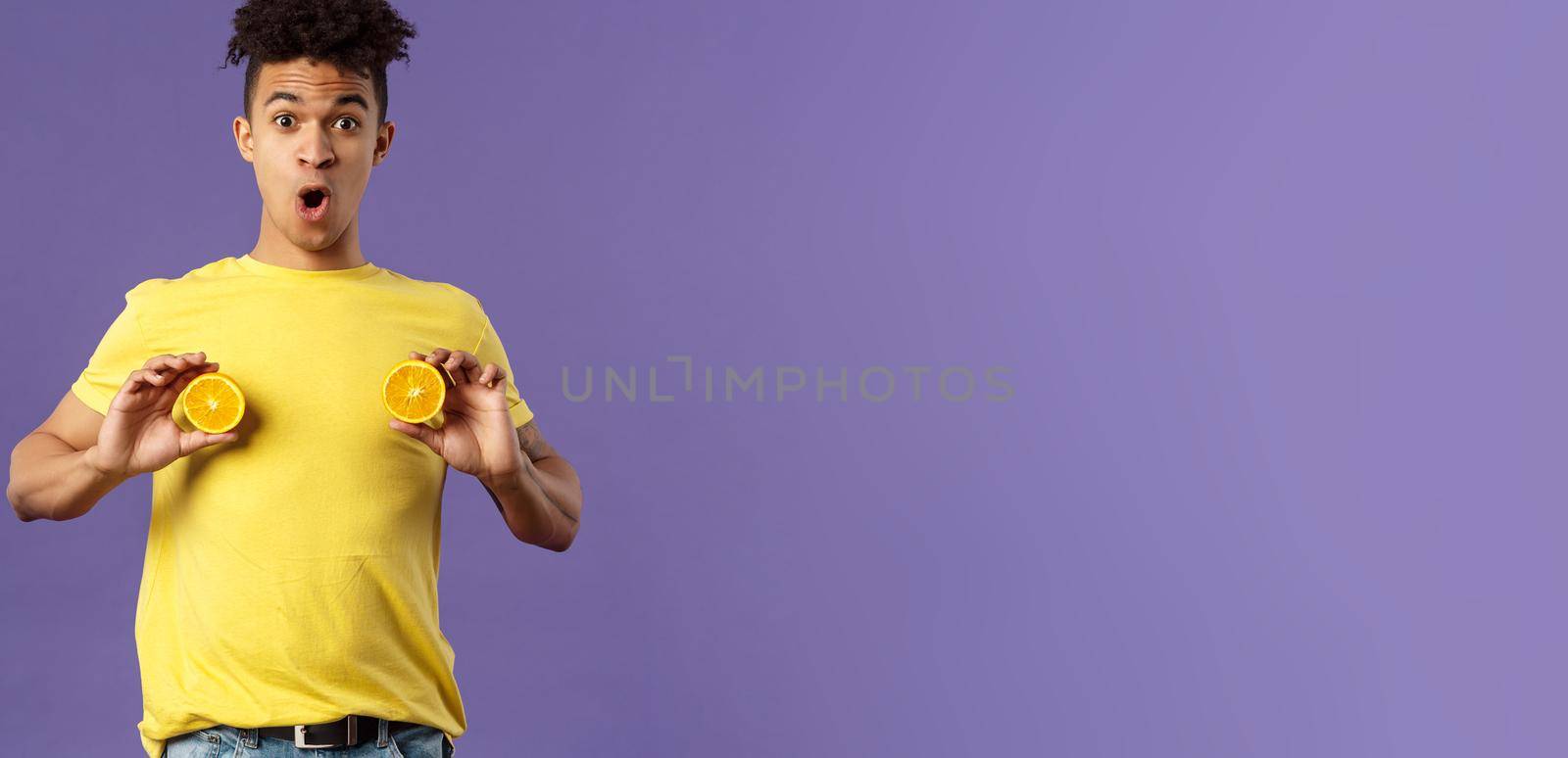 Holidays, vitamins and vacation concept. Portrait of funny and cute young 25s man fool around, showing breast with pieces of oranges over chest, look ashamed or shocked, purple background by Benzoix