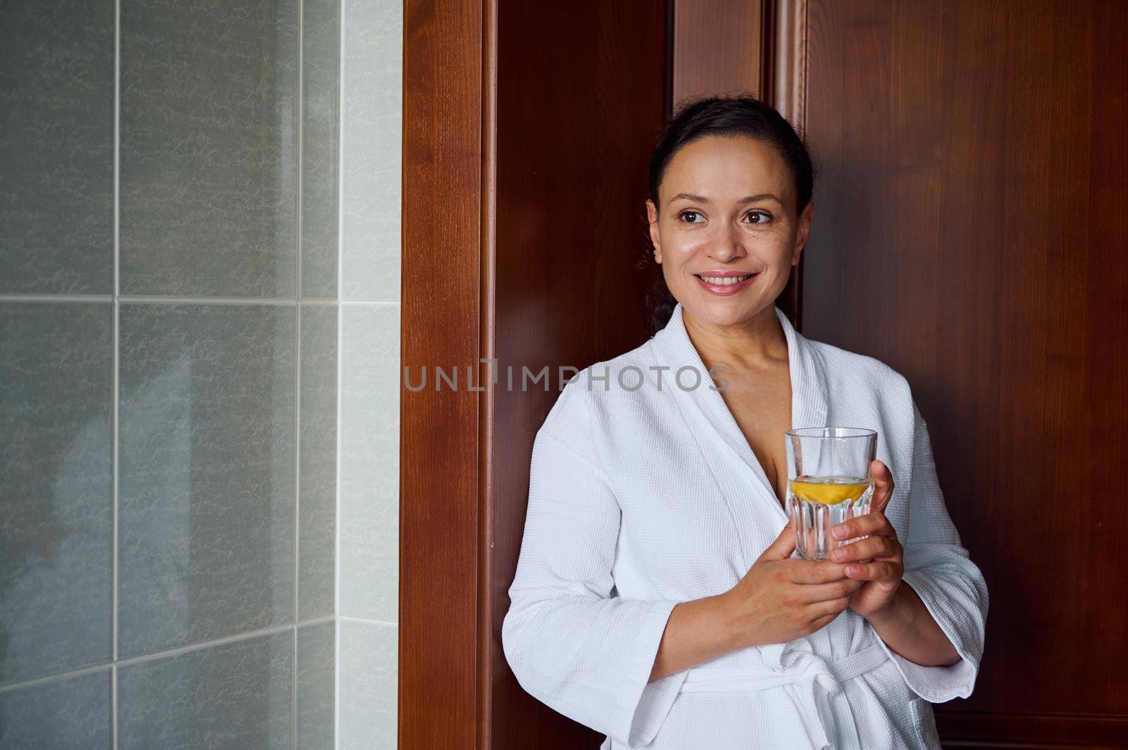 Close-up portrait of a stunning multi-ethnic brunette woman with glowing clean and healthy skin, in bathrobe, holding glass with lemon water for good start to the day. Diet, vitality, healthy habits