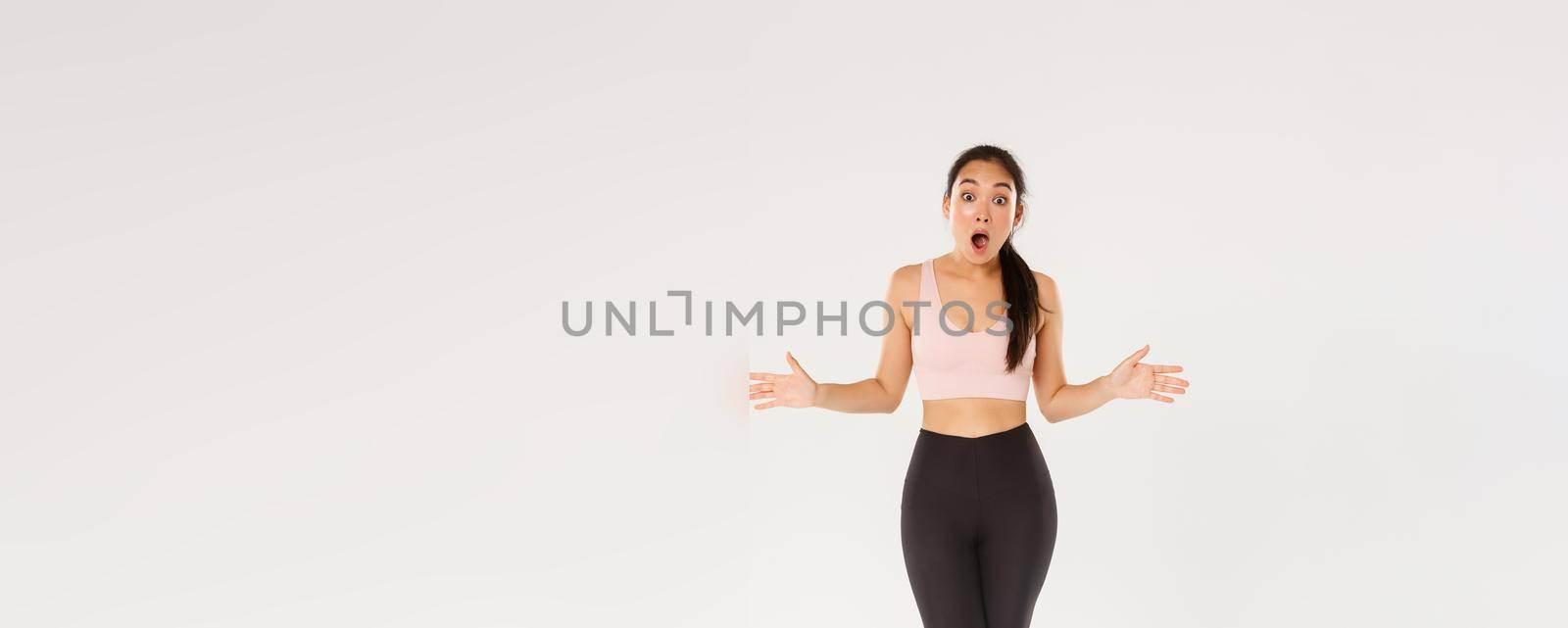 Full length of astonished and surprised female athlete, sportswoman in activewear looking impressed, drop jaw and spread hands sideways see great gym discount, workout equipment in sale.