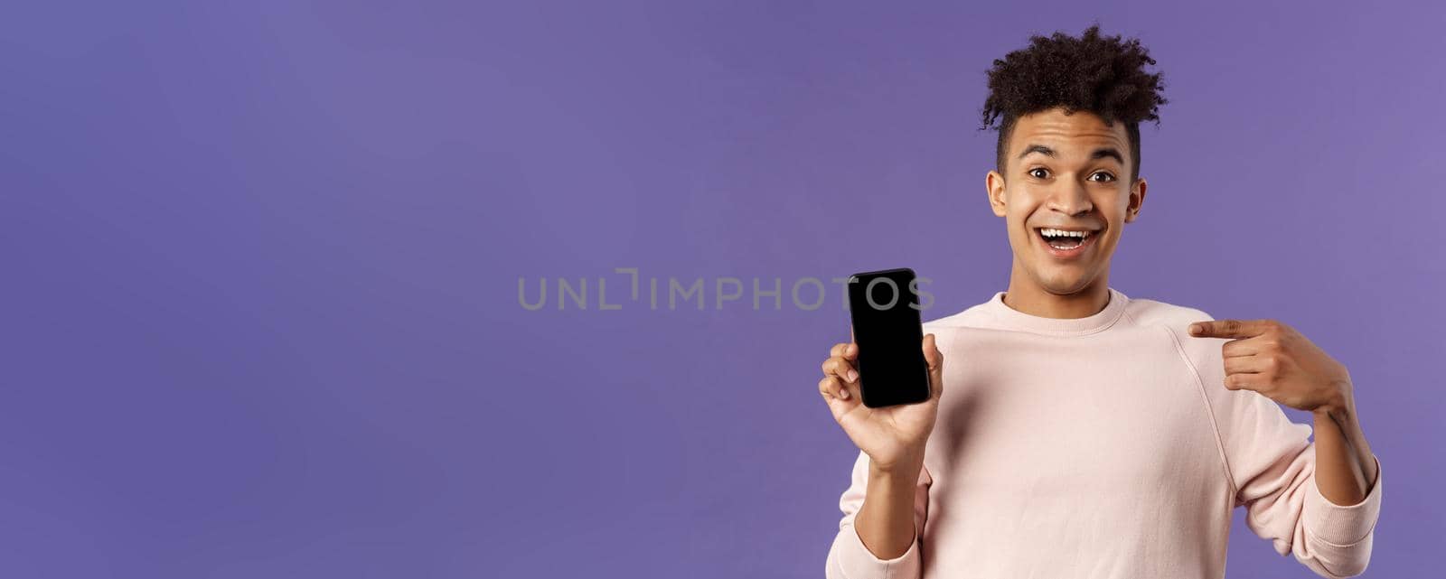 Portrait of excited, happy smiling man talking about new application or game feature, pointing at smartphone display, talking about mobile phone with amused cheerful expression.