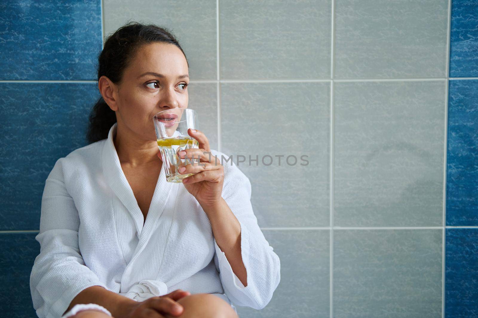 Diet. Vitality. Skin and body care. Healthy habits and lifestyle concept. Beautiful African American woman in white bathrobe, drinking clean lemon water, against bathroom wall background. Copy space