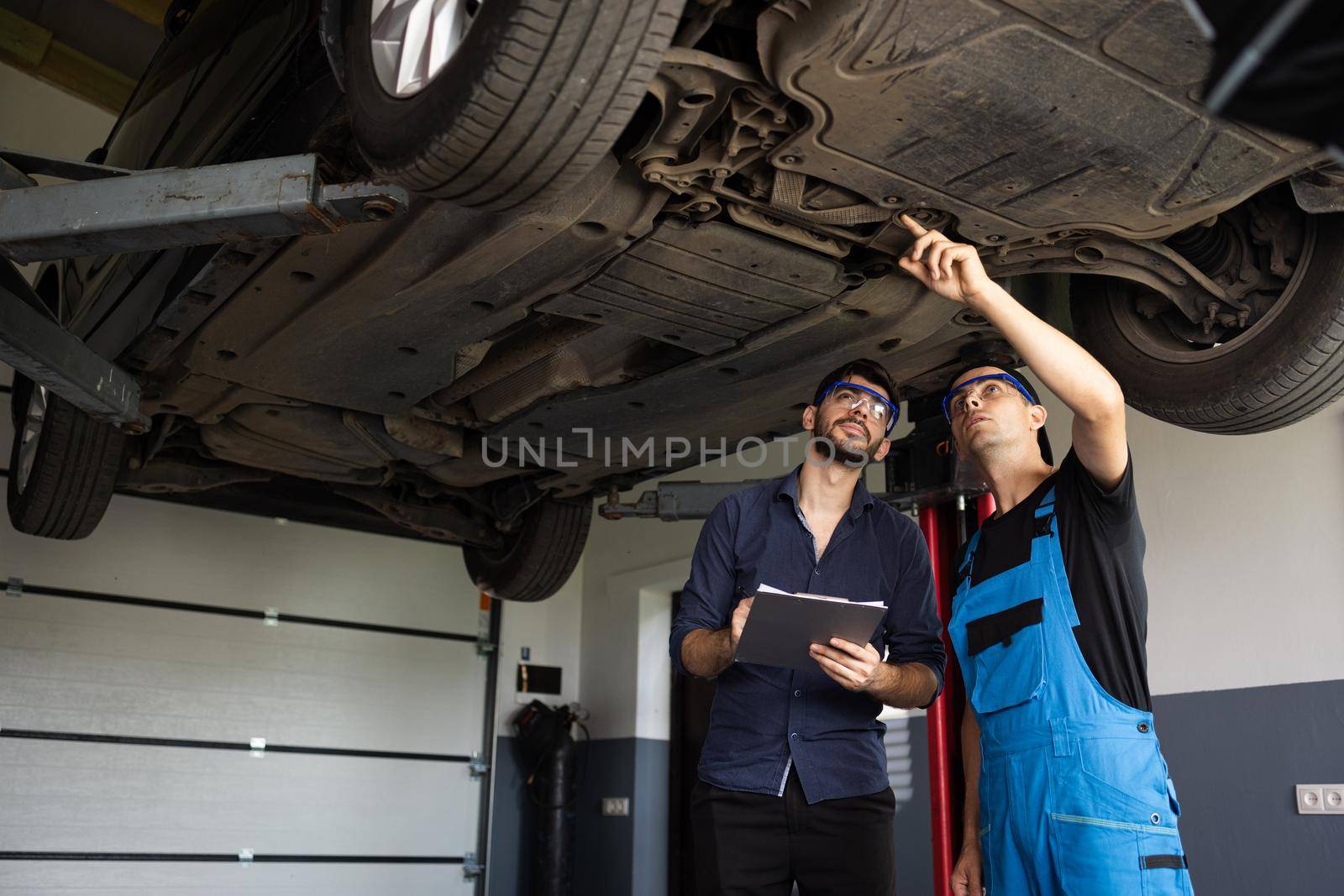 Auto Service. Car Service Employees Inspect the Bottom and Skid Plates of the Car. Manager Checks Data on a Notebook and Explains the Breakdown to a Mechanic. Modern Workshop.
