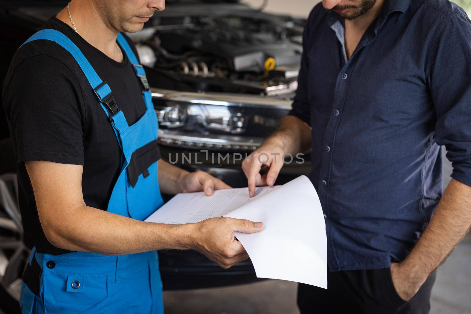 Specialist is showing info on tablet. Car service manager with a tablet, talking to mechanic man discussing car diagnostics and repairing. Mechanic talking to manager near vehicle in car service by uflypro
