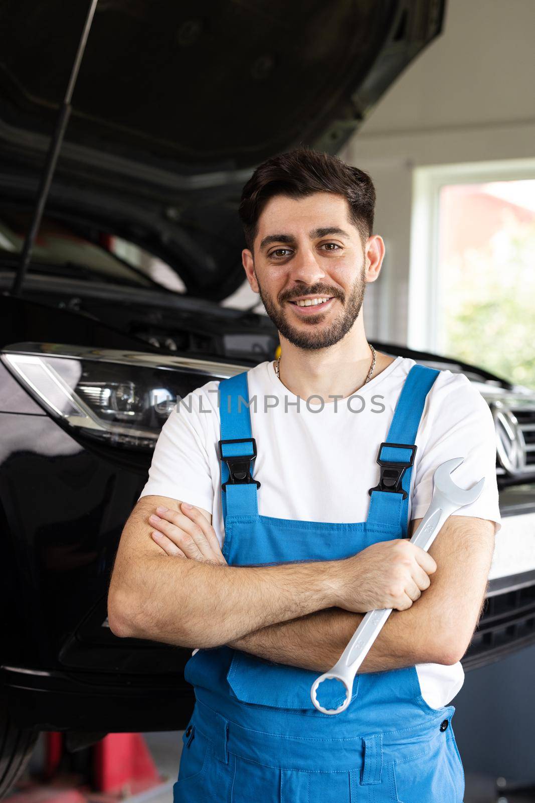 Caucasian bearded man in blue coveralls holds spanner, smiling and looking into camera. Male car mechanic in spacious repair shop by uflypro