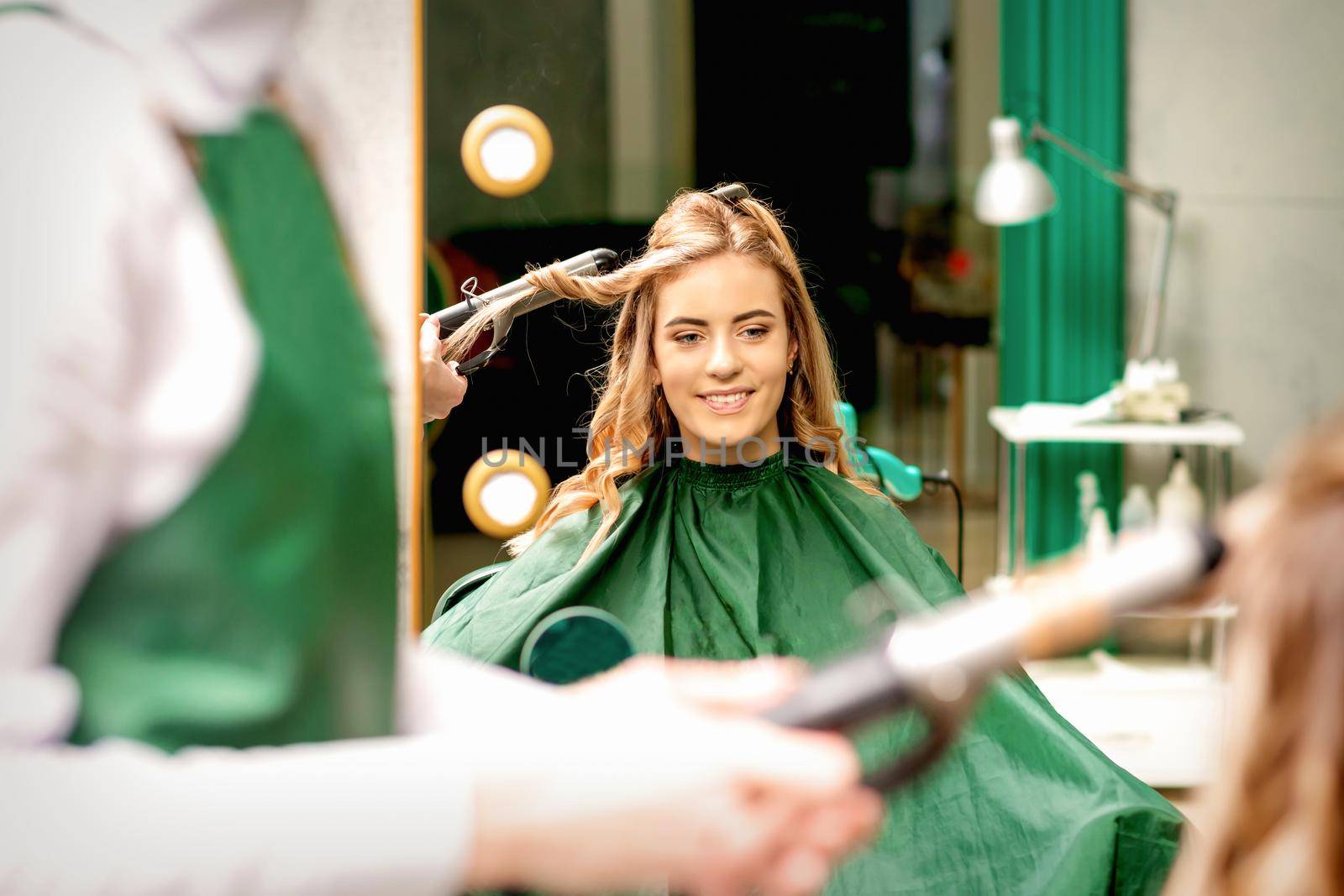Hairdresser using curling tongs curls long brown hair on the young caucasian girl in a beauty salon