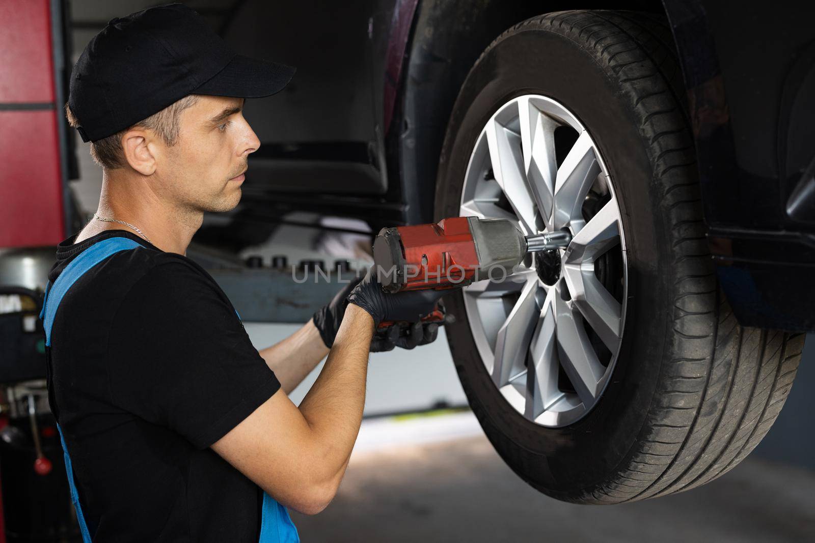 Repairman Works in a Modern Car Service. Specialists Removes the Wheel in Order to Fix a Component on a Vehicle. Mechanic is Unscrewing Lug Nuts with Pneumatic Impact Wrench by uflypro