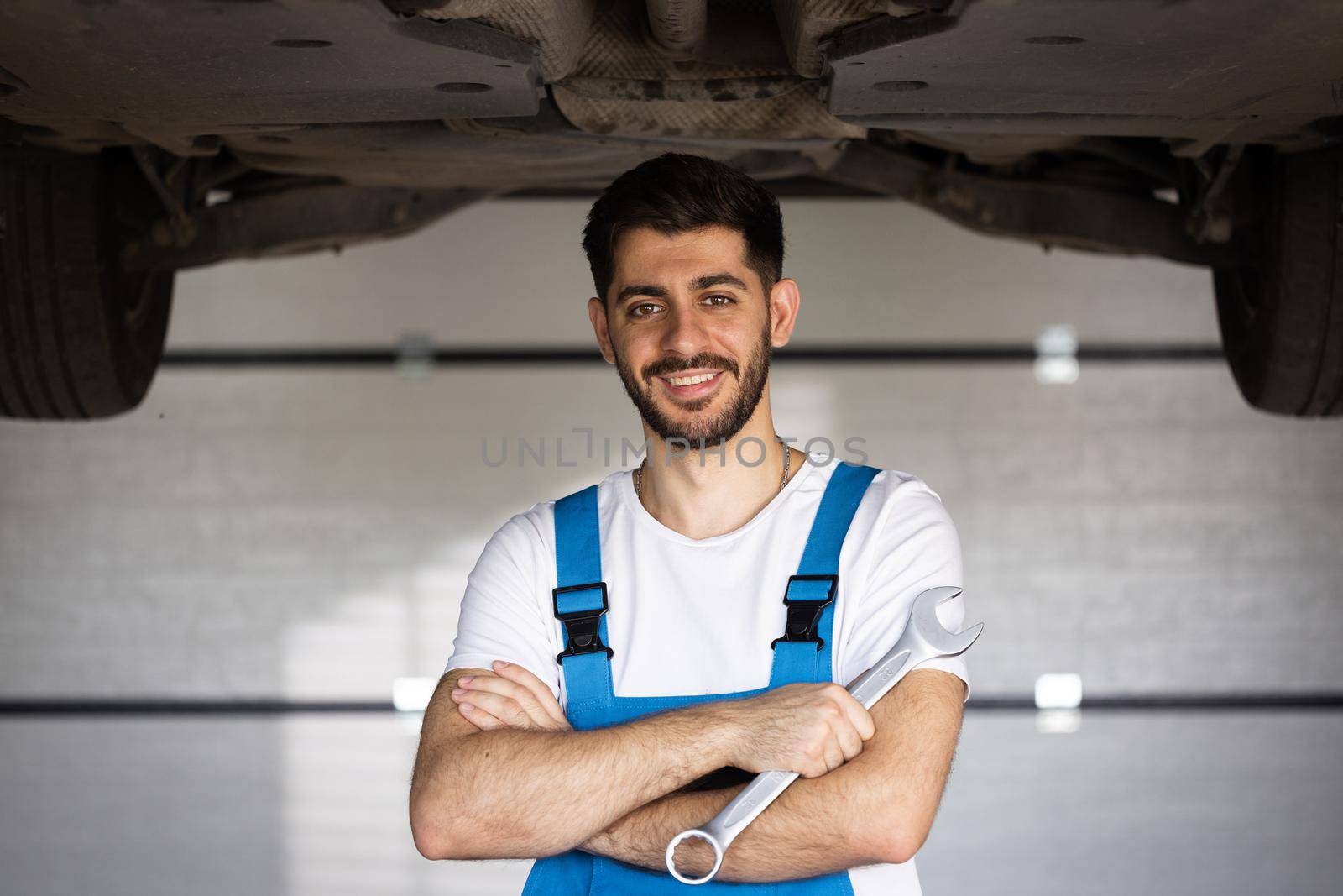 Portrait of bearded mechanic repairing in uniform standing looking camera crossed hands at under lifted car of automobile. Repairing car service concept by uflypro