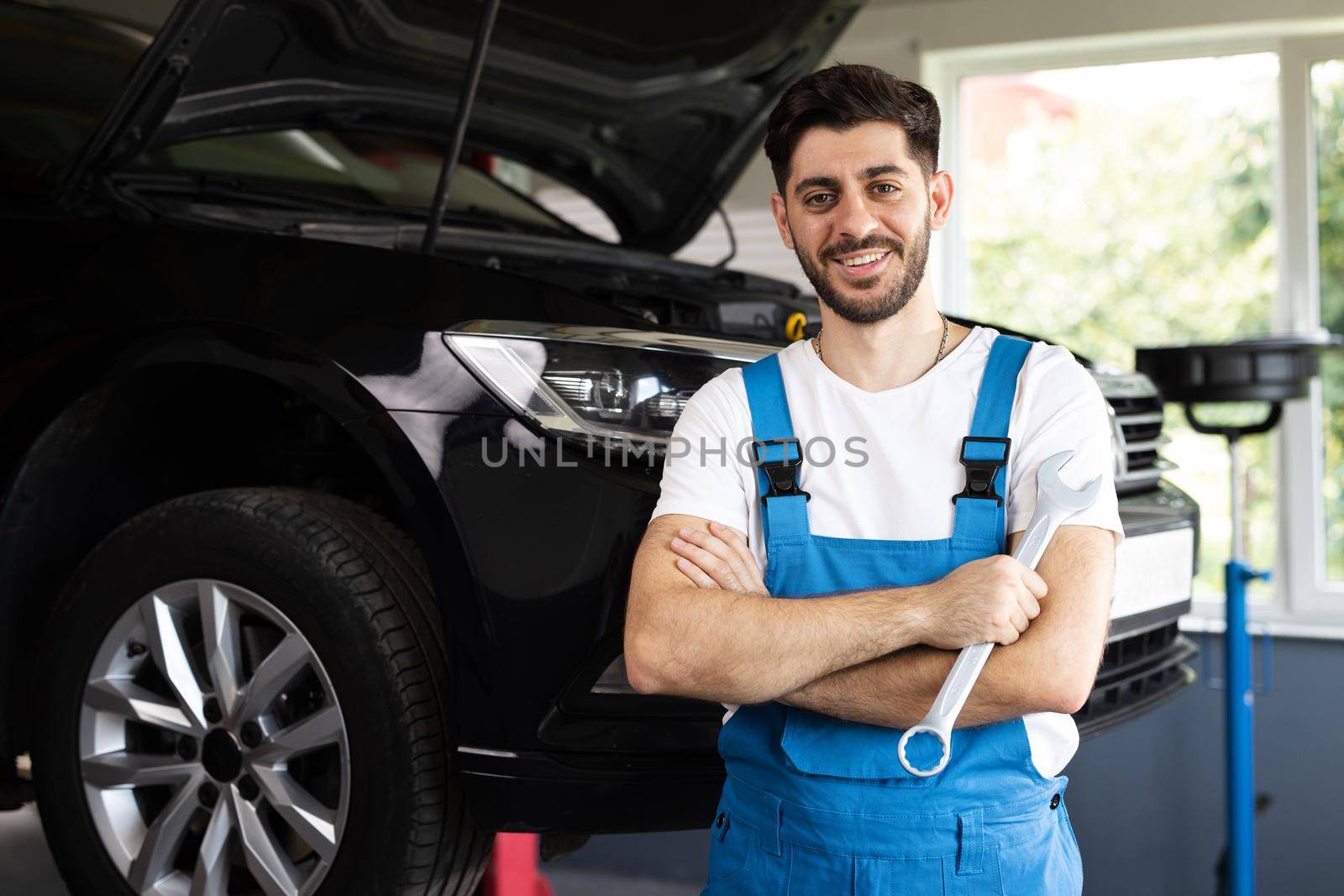 Young caucasian bearded man in blue coveralls holds spanner, smiling and looking into camera. Male car mechanic in spacious repair shop by uflypro