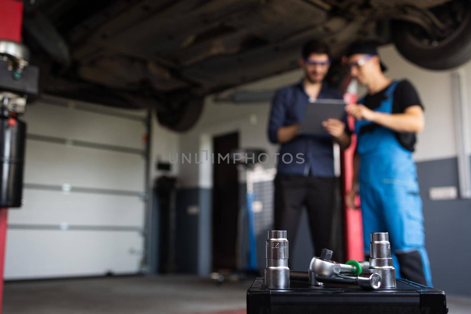 Specialist is Showing Info on a Tablet Computer. Male Mechanic Talking to Manager Under Vehicle in Car Service. Empowering Man in Modern Workshop.