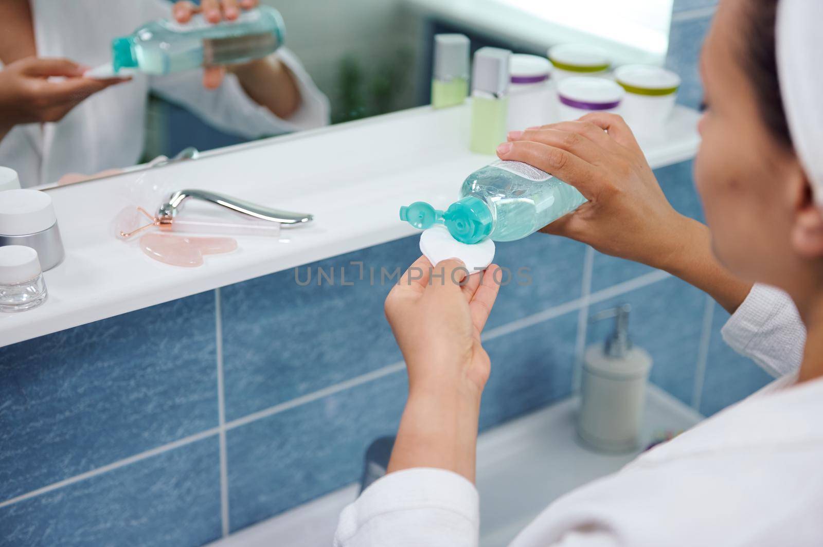 Selective focus on the hands of a blurred woman standing in front of a bathroom mirror and pouring a cleansing lotion on a cotton pad, to clean the face skin and remove make-up. Skin and body care