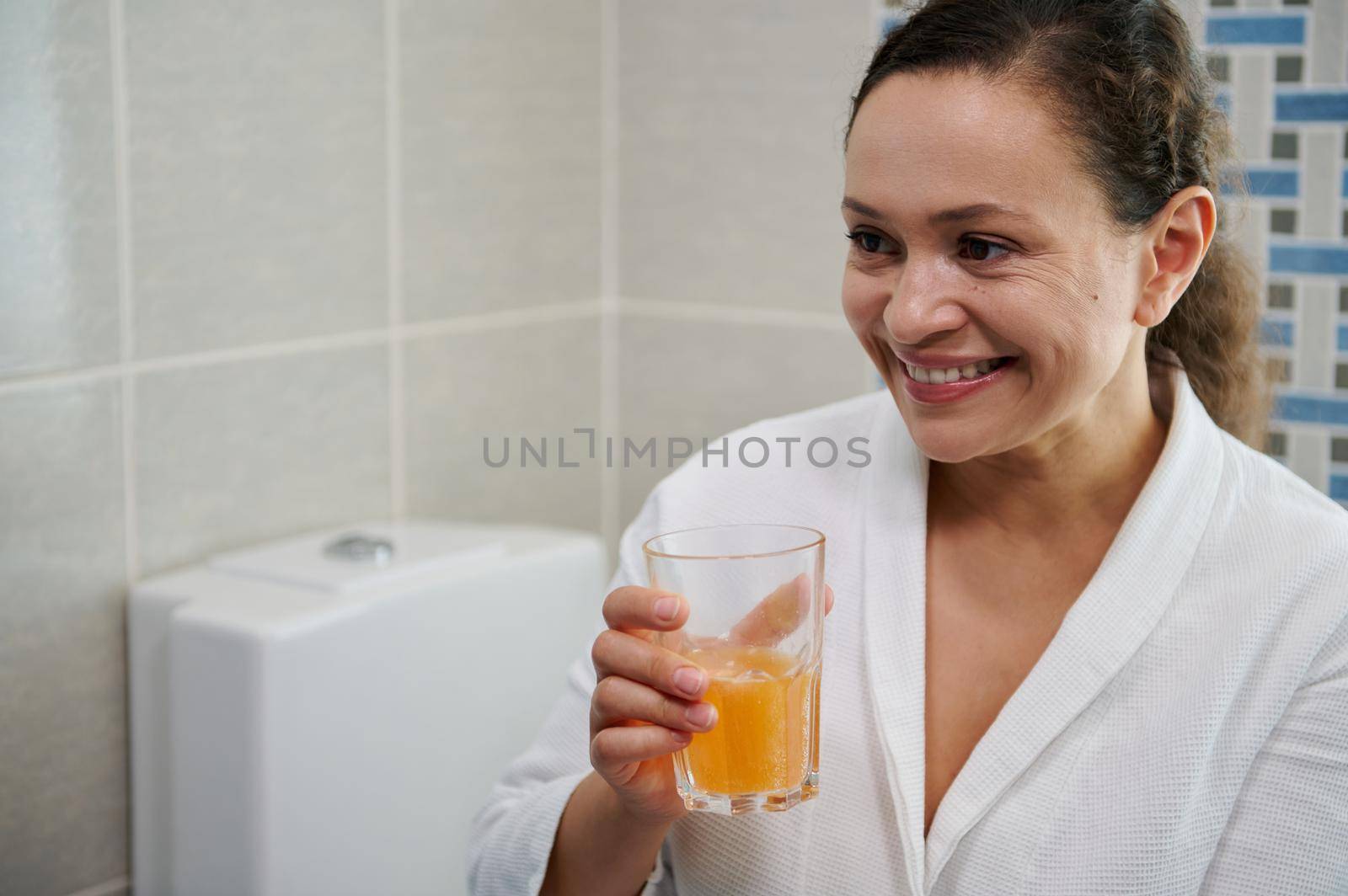 Middle-aged woman in white bathrobe holding a glass of pure water with effervescent tablet, smiling a toothy smile, taking daily dose of vitamins, minerals and dietary nutritional supplement medicine