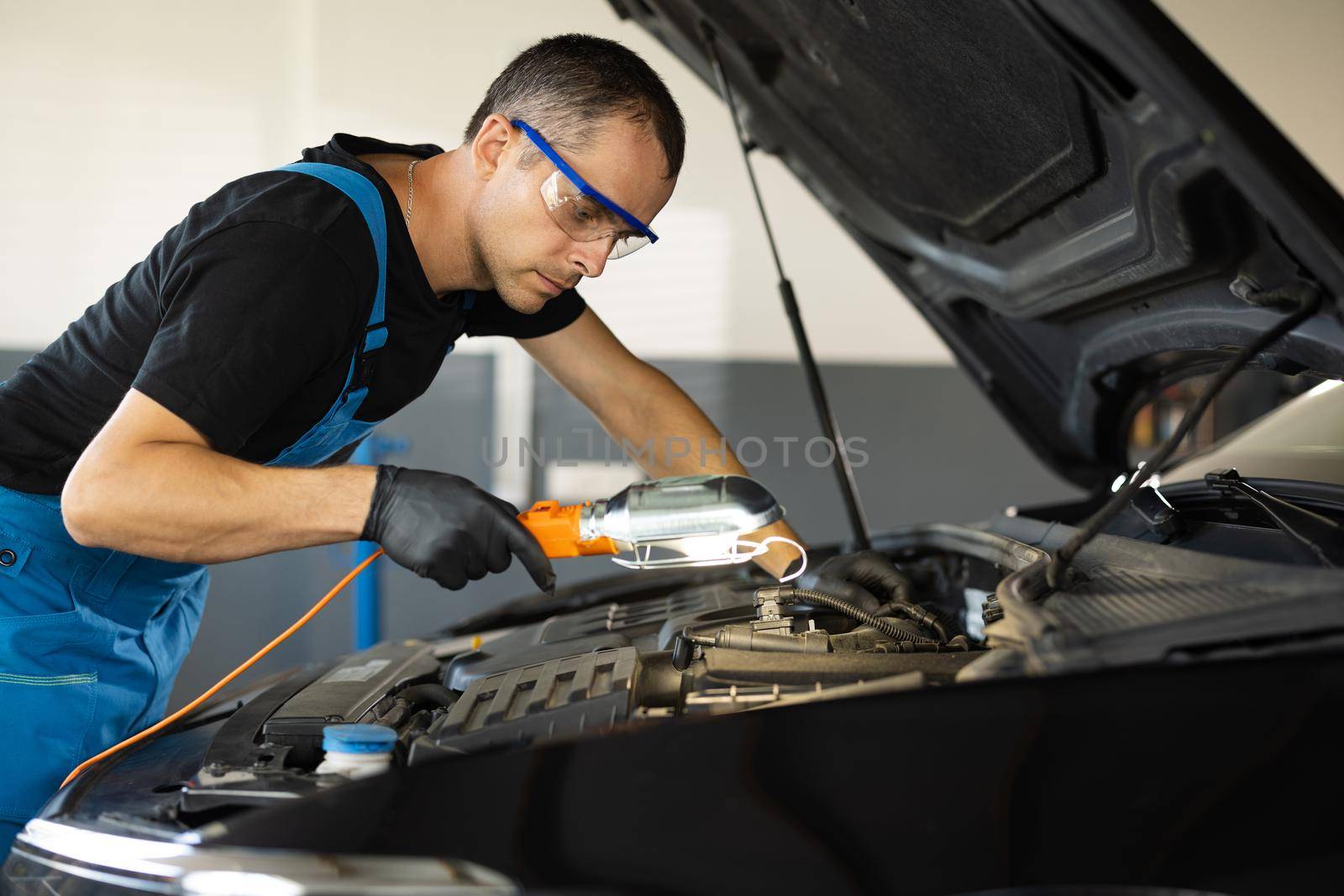 The mechanic in blue overalls and safety glasses inspects the car while working with led lamp. The work of a car mechanic in a modern workshop, car service