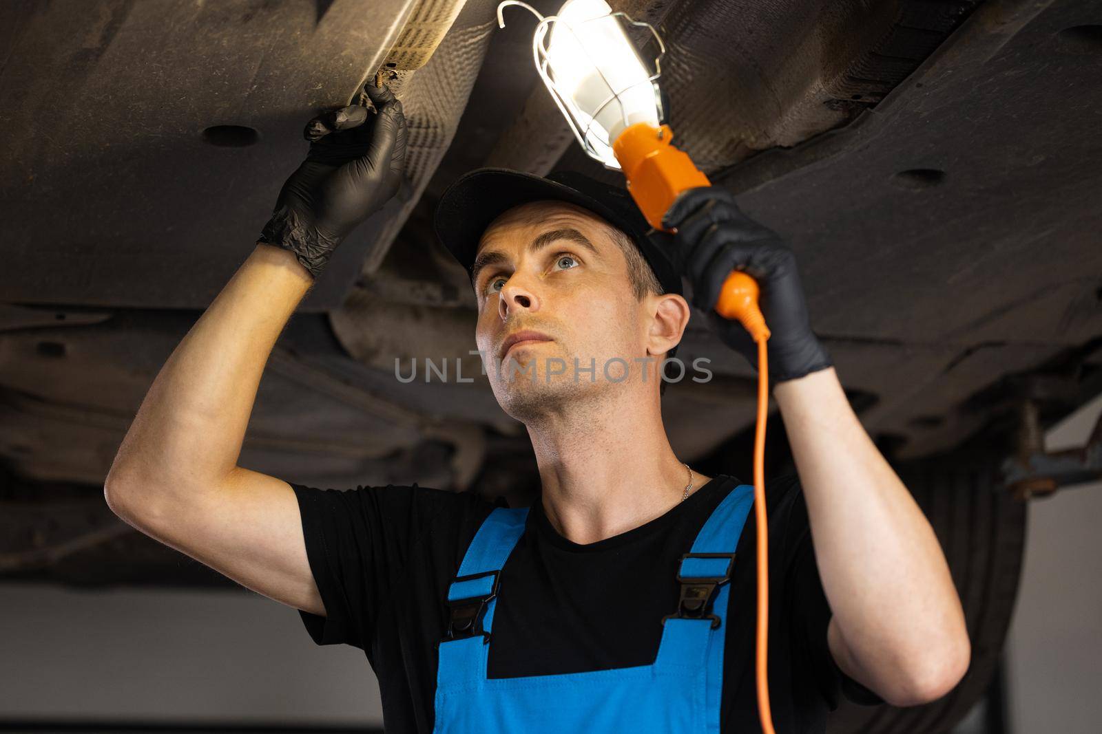Caucasian car mechanic stands with led lamp looking at car parts to wipe the engine under the car on the overhaul to change the oil and maintain the engine in the garage.
