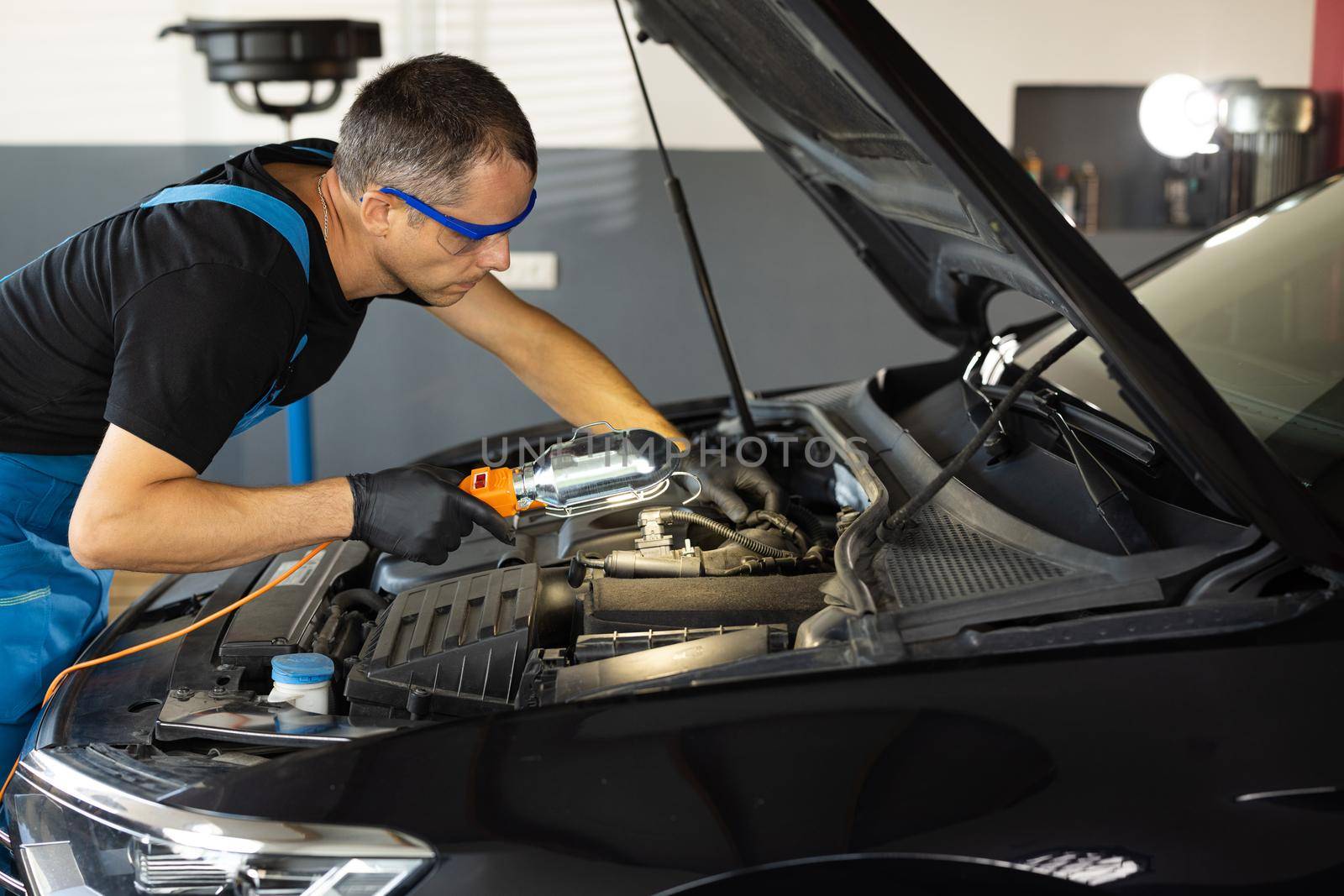 Young caucasian man in blue overalls and safety glasses inspects engine with flashlight. Male car mechanic at work in spacious repair shop. Modern workshop.