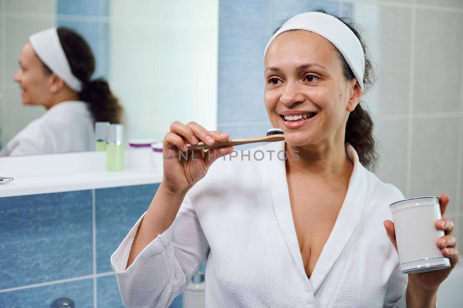 Delightful, natural beauty African American woman wearing white bathrobe, brushing teeth with bamboo eco toothbrush, in the home bathroom. The concept of dental care and oral hygiene. Selective Focus