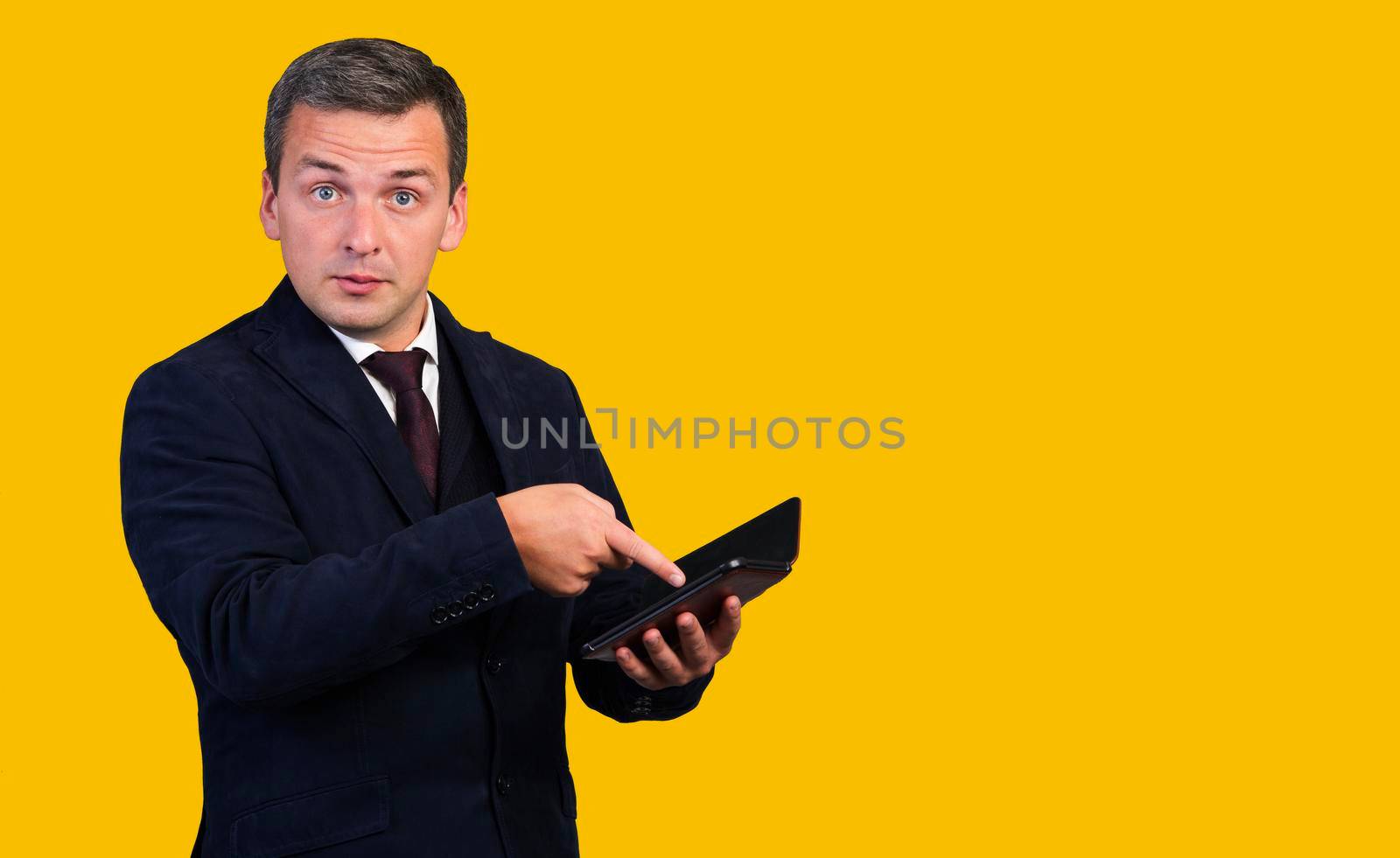 A young man holds a tablet computer in his hands and points his finger at it. . Isolated on yellow background. by gelog67