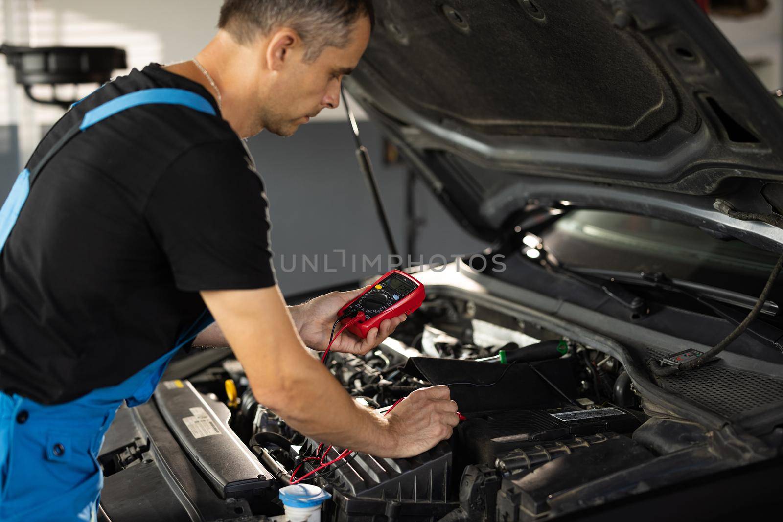 Professional mechanic is working in car service. Good, electrician, electricity, battery charged fully. Check battery voltage with electric multimeter. Car starter battery.