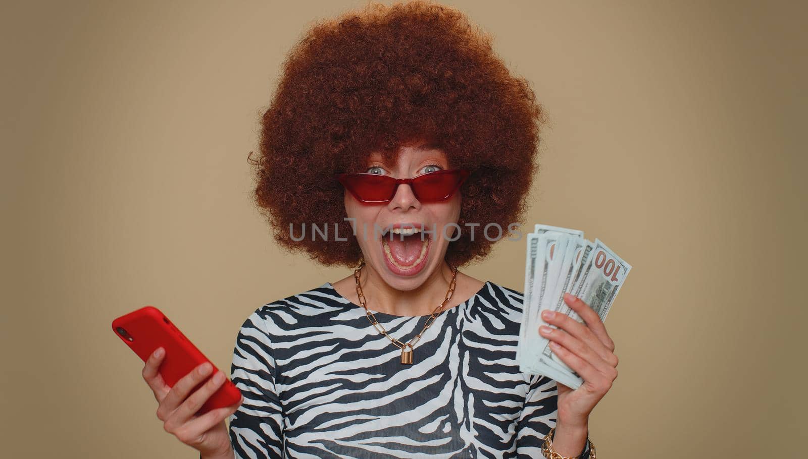 Happy young woman with lush wig looking smartphone display sincerely rejoicing win, receiving money dollar cash banknotes, success lottery luck. Adult stylish female girl on beige studio background