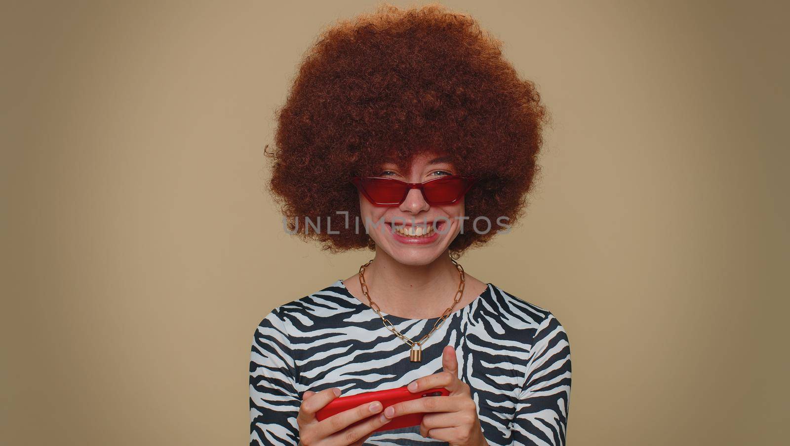 Worried funny young woman with brown lush wig enthusiastically playing racing or shooter video games on smartphone. Adult stylish girl using mobile phone gadget app on beige studio background indoors