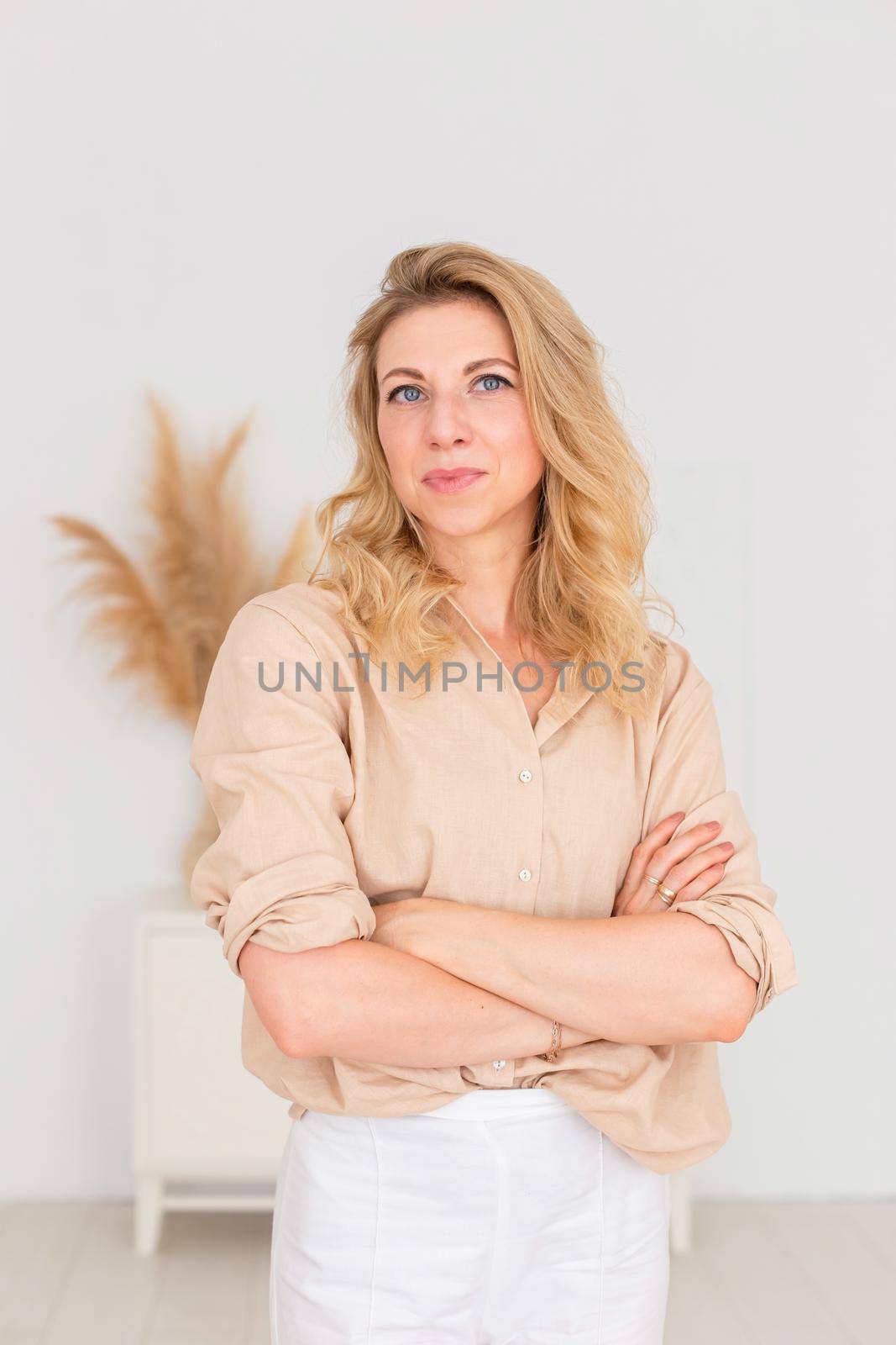 Portrait of a beautiful woman, 30-40 years old, with blond hair, stands in a beige linen shirt and white trousers, in a white room interior. Copy space. Vertical