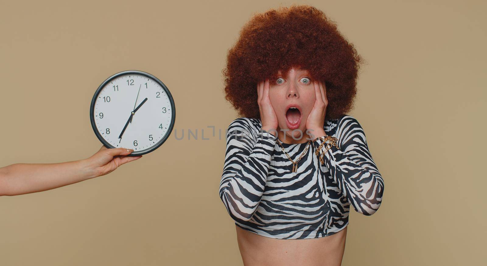 Young woman in brown lush wig with anxiety checking time on clock, running late to work, being in delay, deadline. Adult stylish female girl looking at hour, minutes, worrying to be punctual, indoor