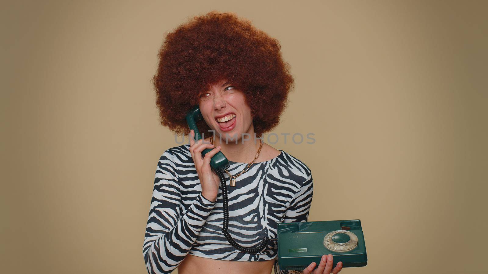Crazy pretty young woman with brown lush wig talking on wired vintage telephone of 80s, fooling, making silly faces. Adult one stylish female girl isolated alone on beige studio background indoors