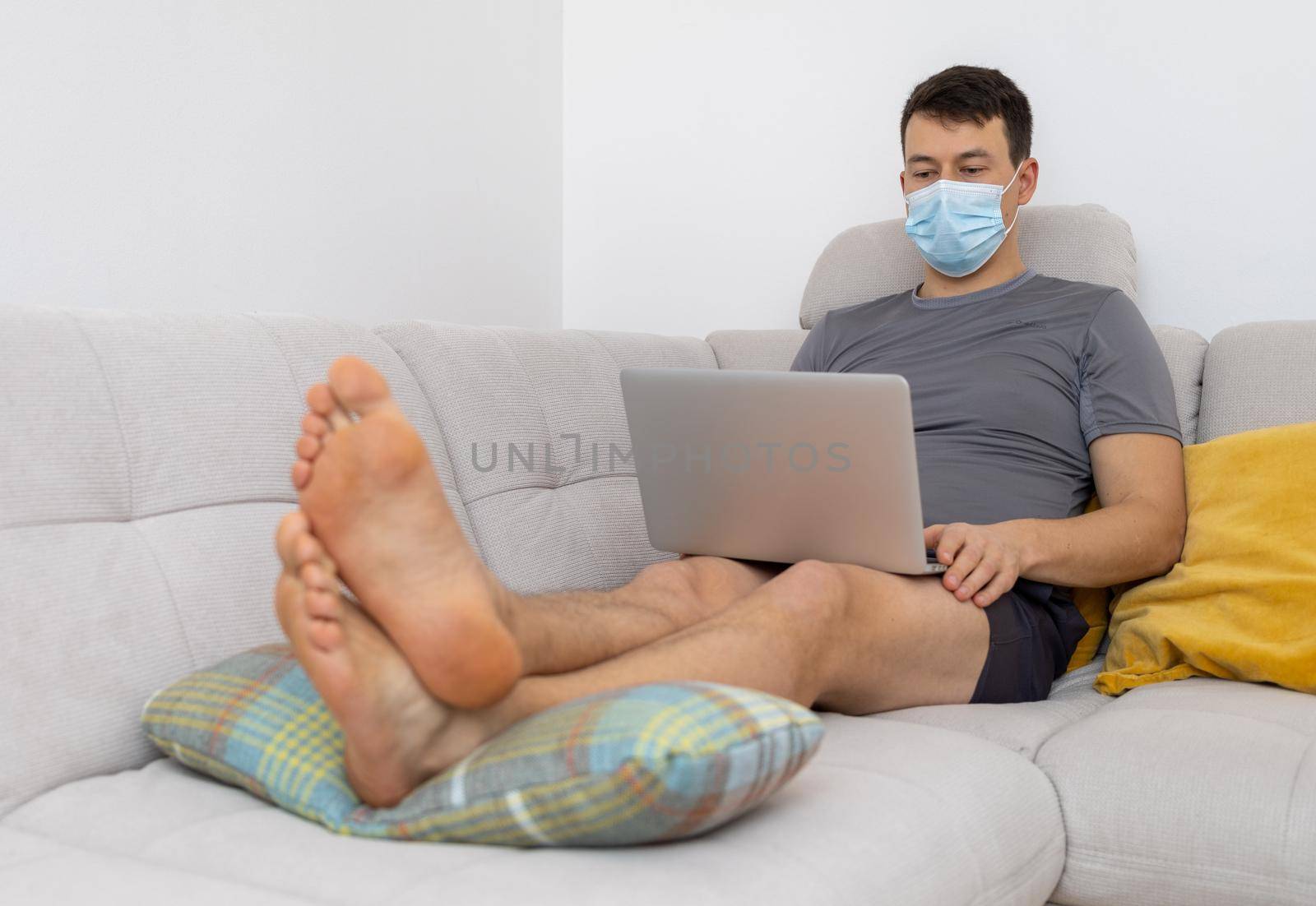 young man in mask and with laptop sitting on sofa by Chechotkin