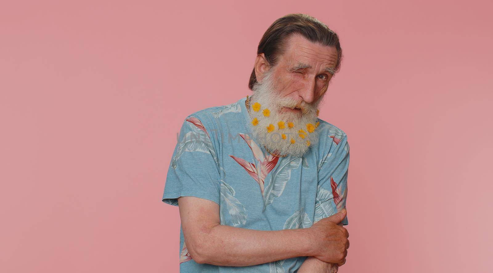 Smiling playful happy senior man with flowers in gray-haired beard blinking eye, looking at camera with toothy smile, winking and flirting, expressing optimism. Elderly grandfather on pink background