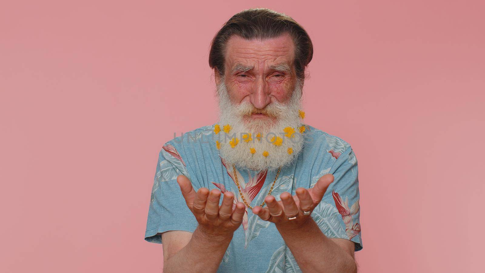 Upset disappointed senior man with flowers in gray-haired beard wipes tears and cries from despair, being sad because of unfair things. Elderly grandfather isolated alone on pink studio background
