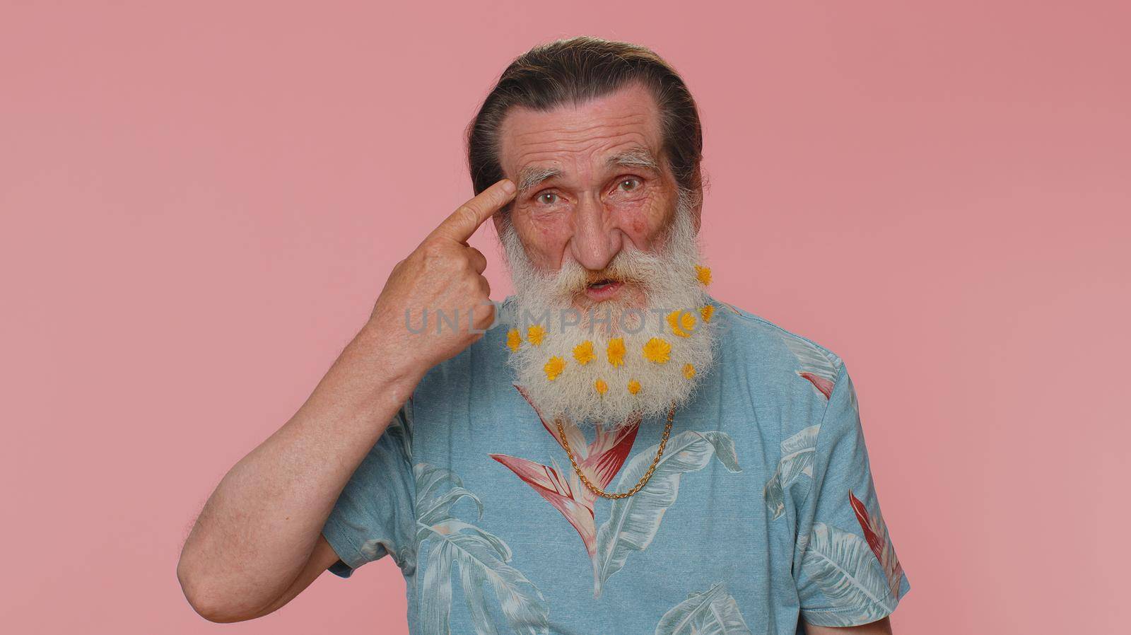 You are crazy, out of mind. Senior man with flowers in beard pointing at camera and showing stupid gesture, blaming some idiot for insane plan. Elderly grandfather alone on pink studio wall background