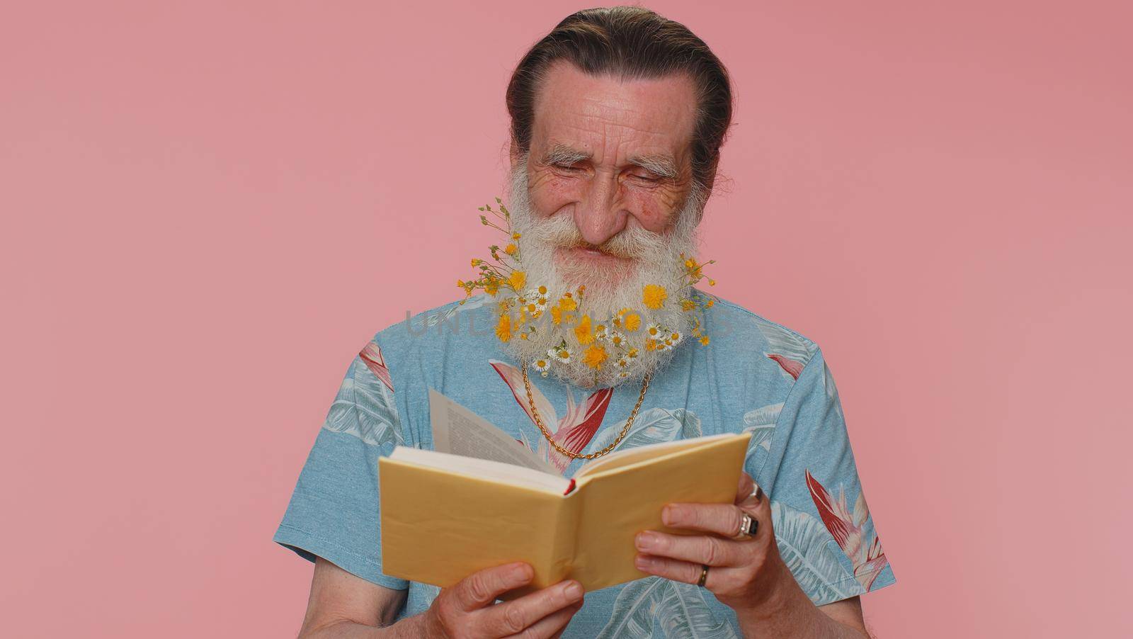 Senior man with flowers in gray-haired beard reading funny interesting fairytale story book, leisure hobby, knowledge wisdom, education, learning, study, wow. Elderly grandfather on pink background