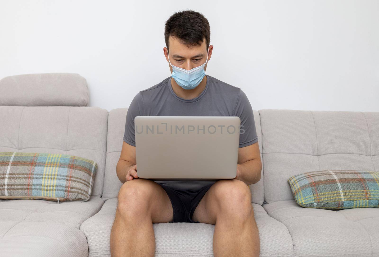 young man in mask and with laptop sitting on sofa by Chechotkin