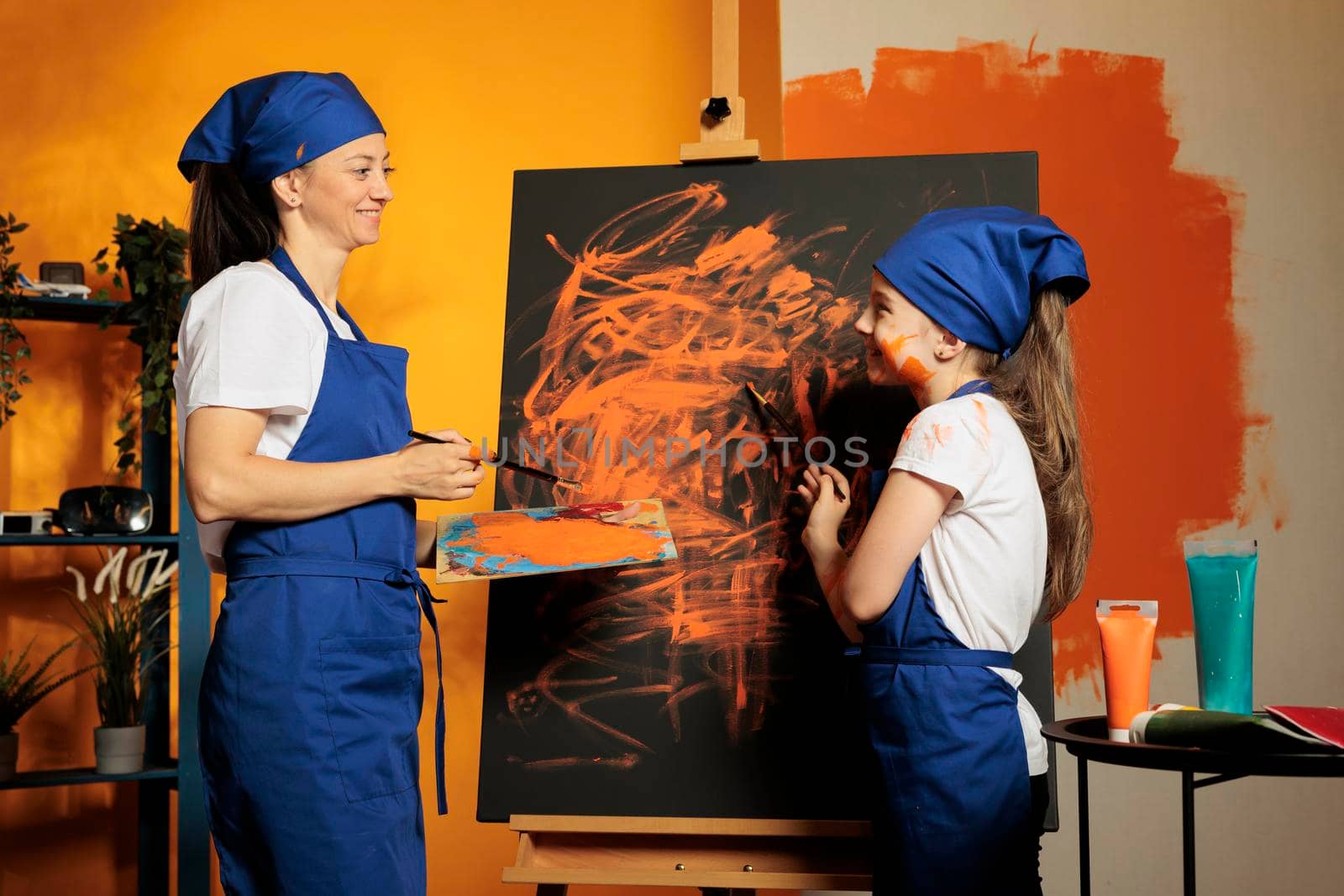Mother and girl using orange paint on canvas, painting artistic masterpiece with watercolor aquarelle, wet paint from palette and brush. Creating colorful artwork with skills and creative vision.