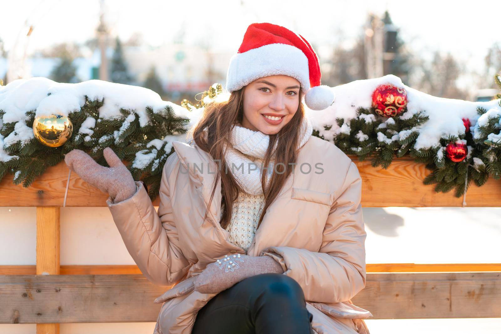 Winter portrait young adult beautiful woman in Santa hat. Christmas mood. Snowing. Winter beauty fashion concept. Pretty young woman sitting bench outdoor near Christmas garland