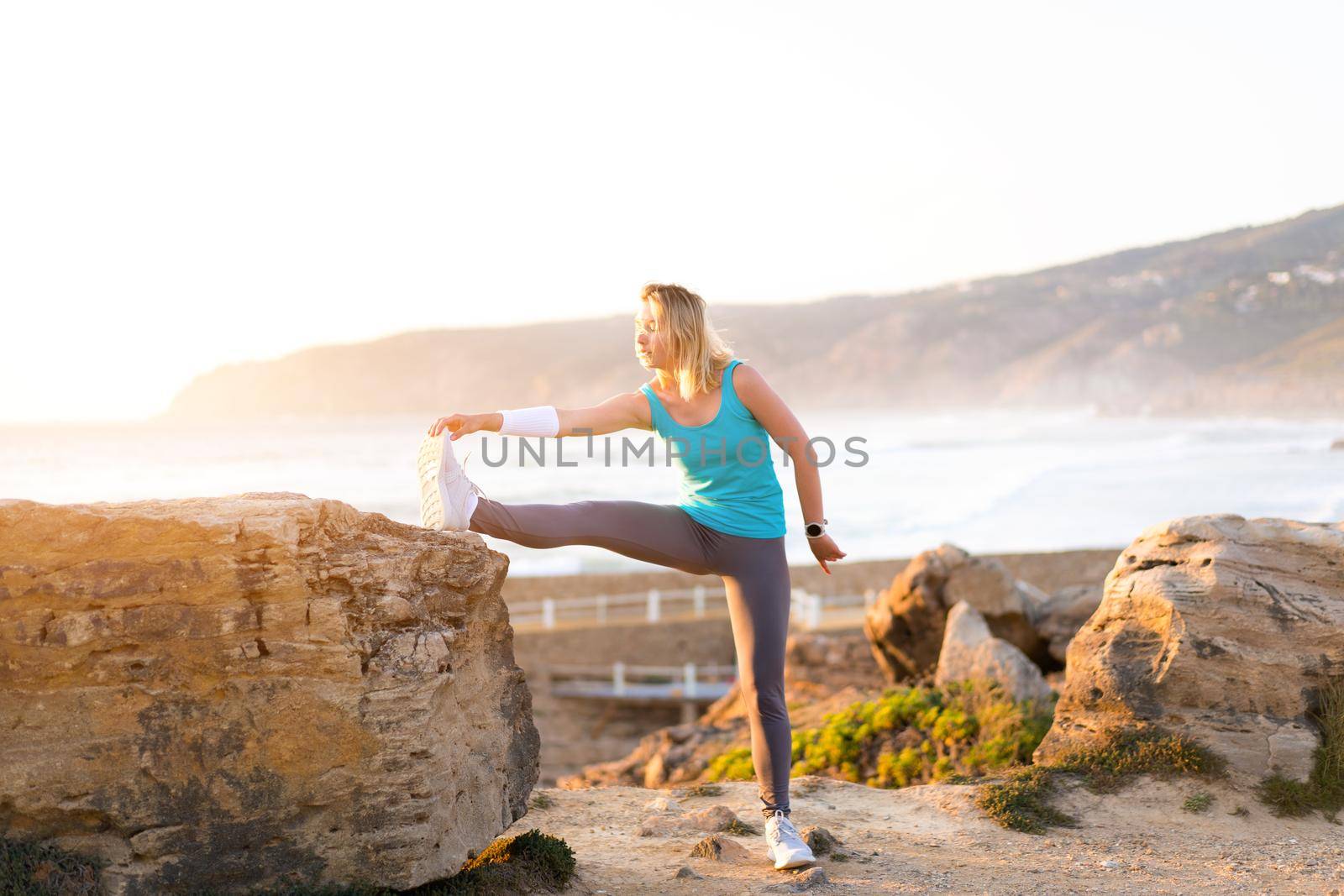 Flexible caucasian girl stretches legs after an intensive running workout. Athletic female middle age warming up before jogging workout outdoor. Dressed sportive wear ocean seashore sunset background