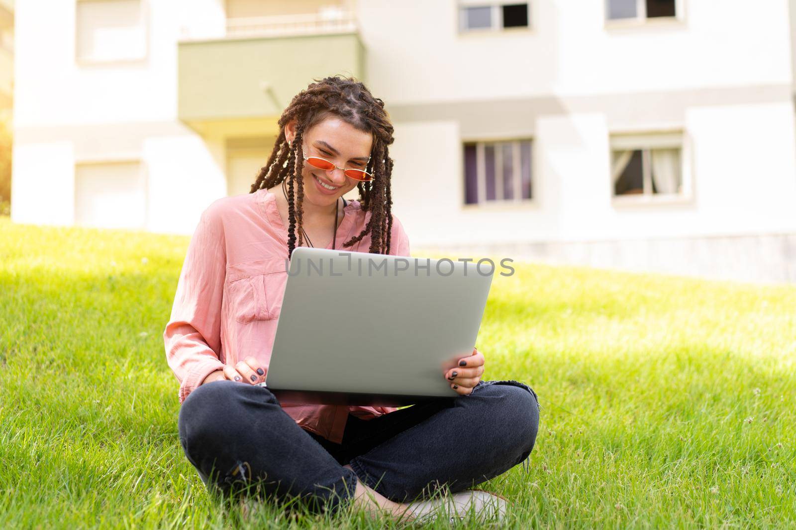 Photo of european woman 25s sitting on green grass in park with legs crossed during summer day while using laptop. Caucasian female hipster with dreadlocks and pink sunglasses on use laptop freelancer