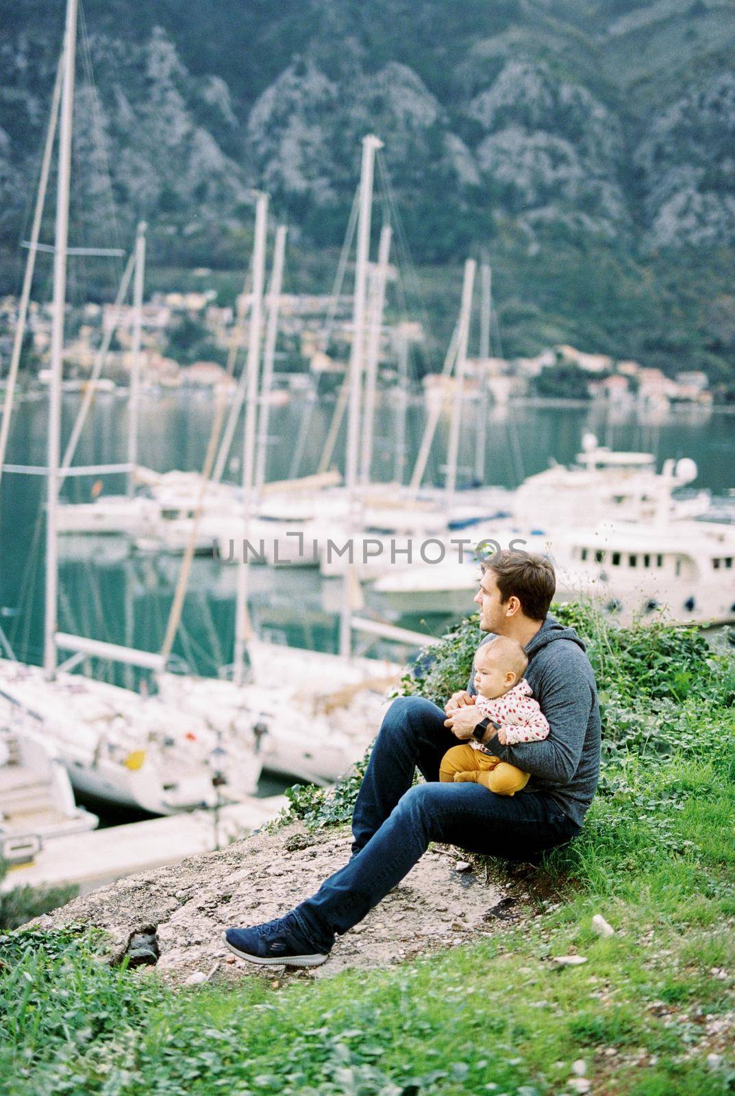 Dad with a baby on his knee sits on a hill and looks at the marina with moored yachts by Nadtochiy
