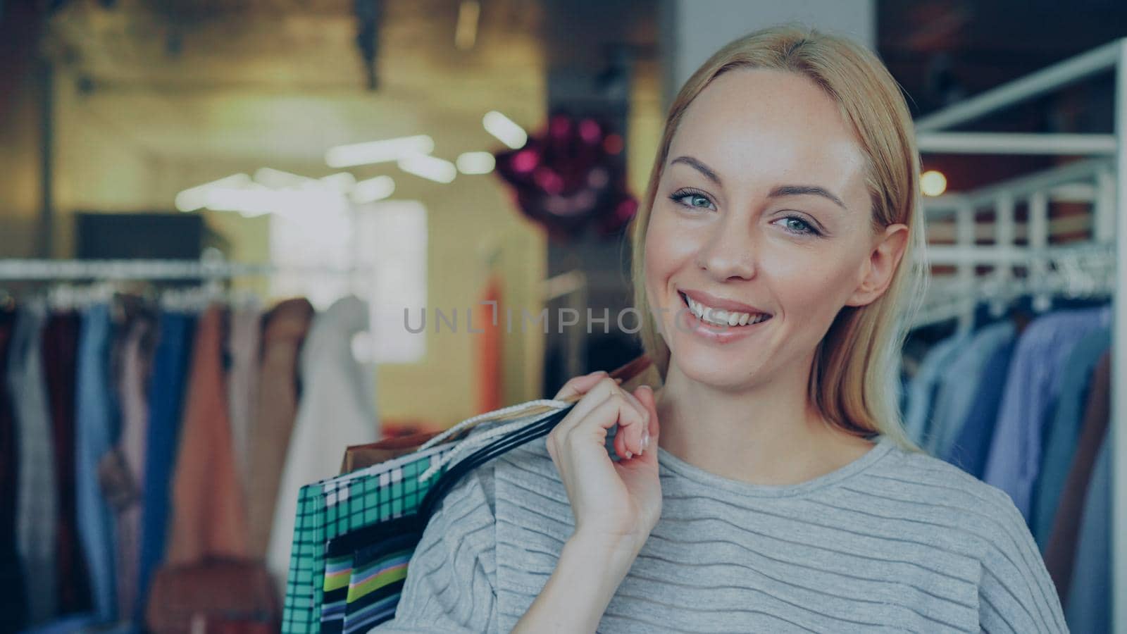 Close-up portrait of blond girl standing with paper bags in clothing boutique, laughing and smiling happily and looking at camera. Stylish clothes is in background. by silverkblack