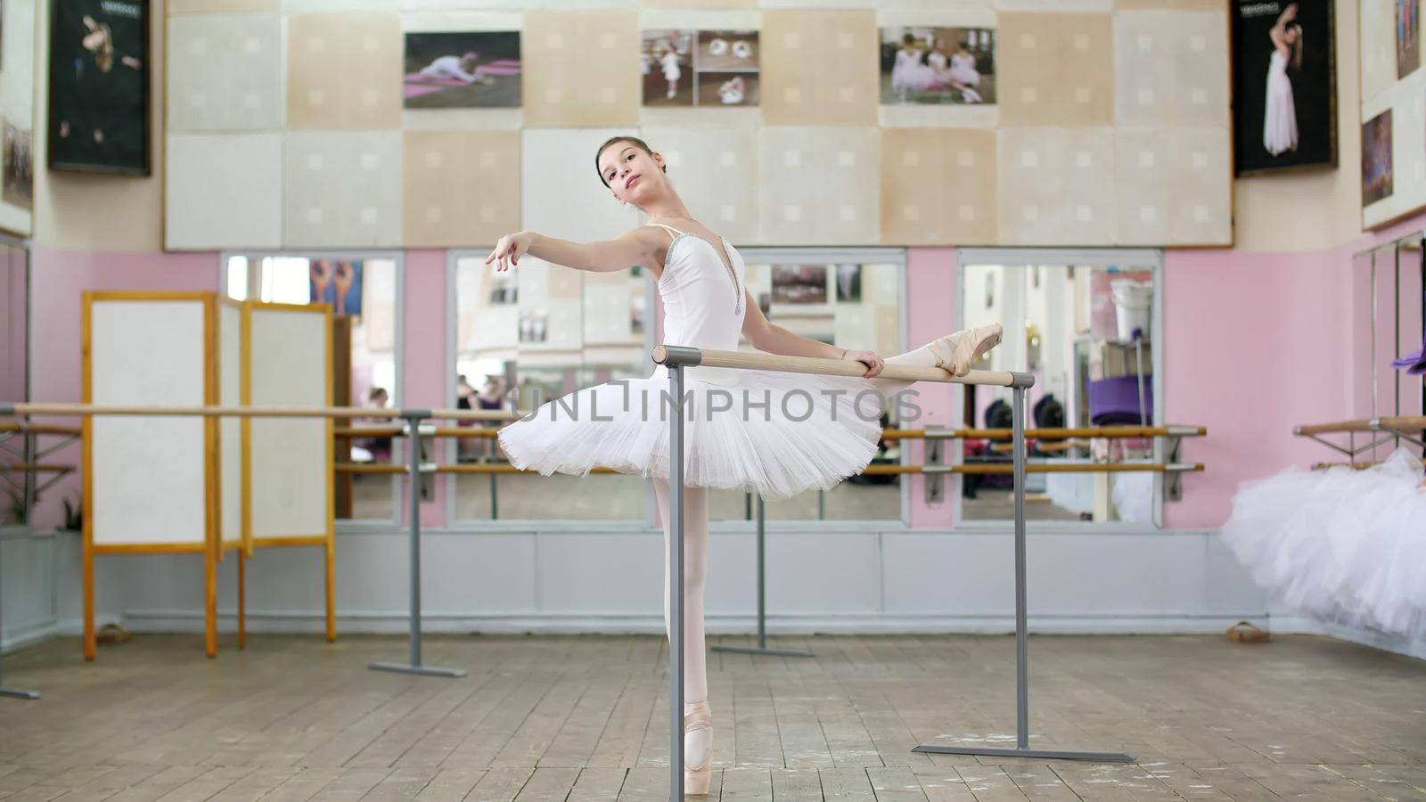 in the ballet hall, girl in white ballet tutu, pack is engaged at ballet, rehearse part de bra with a slope, Raises hand up and down elegantly,Young ballerina standing at railing in hall. High quality photo