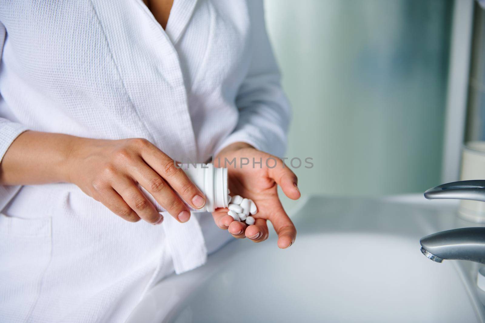 Details: hands of a woman in white bathrobe, holding a container with pills, standing at a sink in bathroom. Patient taking medicine. Compliance to doctor's prescription. Healthy lifestyle. Close-up